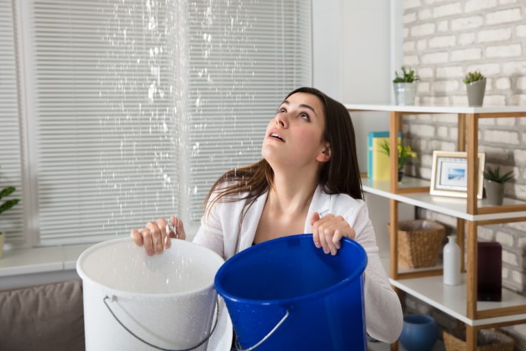 Woman holding blue and white buckets catching water from a leaking roof.