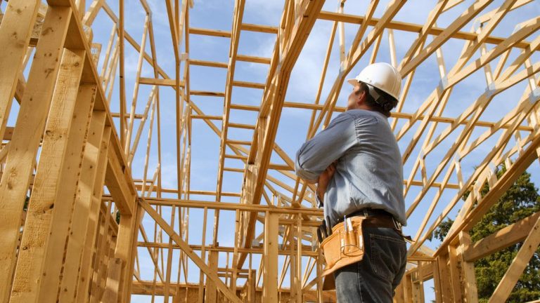 Tradesman wearing a hard head and tool belt on a construction site looking up at the timber frame of a building.